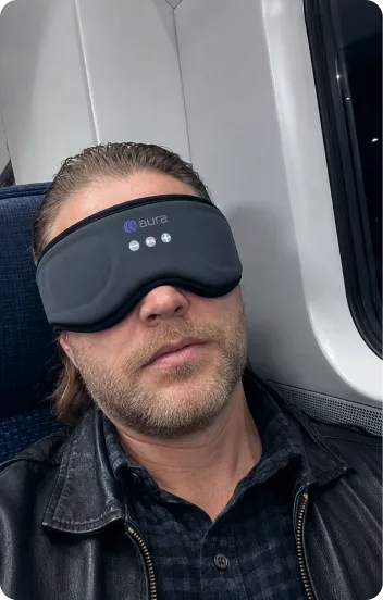 Traveler napping upright in a train seat with the AURA Sleep Mask blocking the cabin light