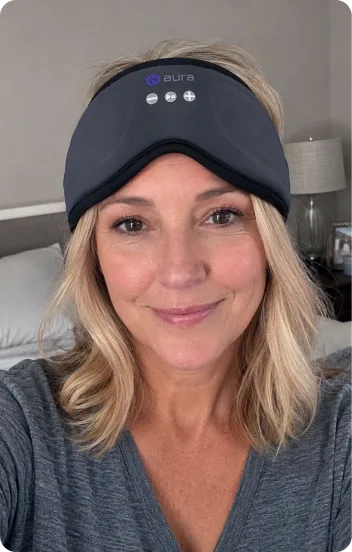 Smiling woman in her bedroom wearing the AURA Bluetooth Sleep Mask pushed up on her forehead